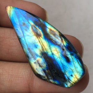 Puede incluir: Una piedra preciosa de labradorita pulida con una vibrante exhibición de colores iridiscentes. La piedra presenta una mezcla de azules, verdes y amarillos, con inclusiones de color gris oscuro y negro. La forma es alargada e irregular.