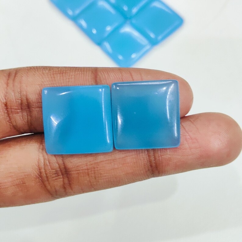 May include: Two square, light blue gemstones with rounded corners are held in a hand. Additional blue square gemstones are visible in the background. The gemstones have a smooth, polished surface.