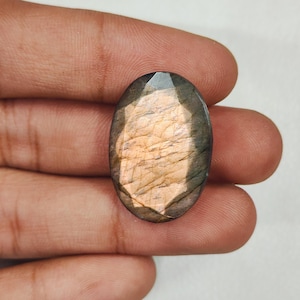May include: An oval-shaped labradorite gemstone featuring a faceted surface with a prominent peach and orange iridescent flash at the centre, surrounded by dark grey and brown tones that transition towards the edges of the stone.