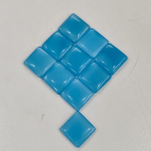 May include: A diamond arrangement of light blue, square glass tiles. Each tile has rounded edges and a glossy finish, reflecting light. The arrangement includes a single tile at the bottom. The tiles are approximately 2.5 cm square.