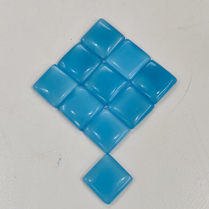 May include: A diamond arrangement of light blue, square glass tiles. Each tile has rounded edges and a glossy finish, reflecting light. The arrangement includes a single tile at the bottom. The tiles are approximately 2.5 cm square.