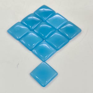 May include: Eleven light blue, square glass tiles. Ten are arranged in a diamond pattern, with one tile below. The tiles have a glossy finish, possibly for crafts or decoration. Each tile is approximately 2.5 cm x 2.5 cm.