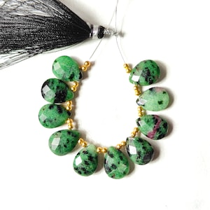 Könnte beinhalten: Strang aus grünen und schwarzen Rubin-Zoisit-Edelsteinperlen. Die tropfenförmigen Perlen sind facettiert und mit goldfarbenen Abstandshaltern aufgereiht. Am oberen Ende des Strangs ist eine schwarze Quaste befestigt.