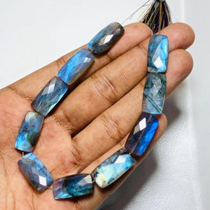 Pode incluir: Um fio de contas facetadas de labradorite. As contas retangulares têm um acabamento polido e iridescente, exibindo tons de azul, verde e cinza. As contas são enfiadas, prontas para a criação de joias. O fundo é branco neutro.