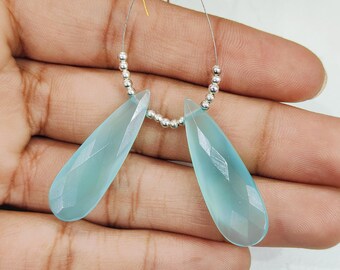 May include: Two light blue teardrop-shaped gemstones with faceted surfaces, strung together with a silver-colored wire and small silver beads. The gemstones are a soft, translucent color, and the overall presentation is delicate and elegant.