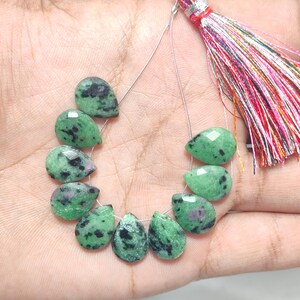 Puede incluir: Un hilo de cuentas de piedras preciosas verdes y negras en forma de lágrima. Las cuentas están ensartadas en un alambre fino, con una borla colorida en la parte superior. Las piedras preciosas tienen un aspecto moteado, con una mezcla de colores verdes y negros.