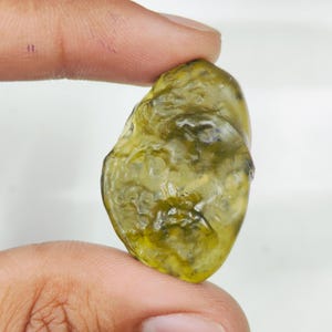 May include: A translucent, olive-green gemstone with an irregular, textured surface. The stone displays varying shades of green, ranging from light yellow-green to deeper forest green tones, with a glossy, uneven finish across its entire shape.