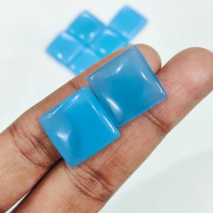 May include: Two square, sky-blue gemstones are held in a hand, with several more in the background. The gemstones have rounded corners and a glossy finish. The gems are a light, bright blue colour.