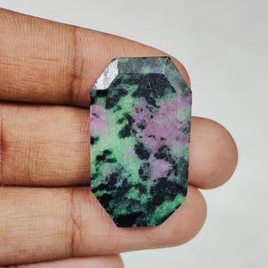 Può includere: Una gemma rettangolare con una parte superiore sfaccettata. La pietra presenta un mix di colori, tra cui verde, rosa e nero. La gemma è tenuta in una mano.