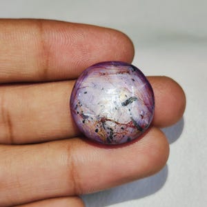 May include: A round, cabochon-cut gemstone featuring a translucent lavender and light purple base with visible internal inclusions, including dark black specks and thin, reddish-brown vein-like patterns throughout the polished surface.