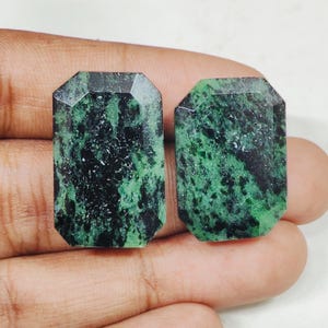 Puede incluir: Dos piedras preciosas de talla esmeralda con un patrón moteado verde oscuro y negro. Las gemas tienen una parte superior facetada y una forma rectangular. Las piedras se sostienen en una mano, mostrando sus superficies pulidas y variaciones de color naturales.
