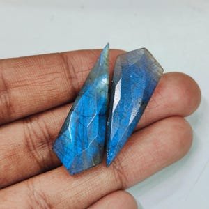 Può includere: Due pietre preziose di labradorite sfaccettate con una combinazione di colori blu e grigio. Le pietre hanno un taglio geometrico con molteplici sfaccettature, creando un effetto scintillante. Le pietre sono tenute in una mano.