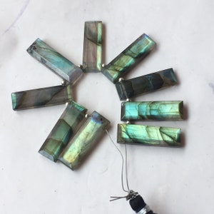 May include: A collection of rectangular labradorite gemstone beads. The beads are a mix of gray, blue, and green, with a faceted cut. Each bead has a small silver bead at each end, strung on a wire.