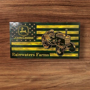 May include: A wooden sign with a green and yellow American flag design. The flag features a John Deere tractor and the words "Rainwaters Farms" in gold lettering.