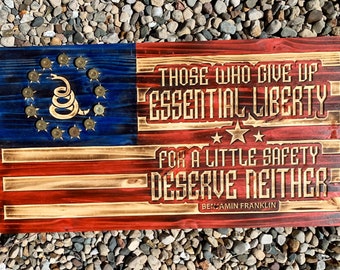 Op de afbeelding: Een Amerikaanse vlag van hout met een rood, wit en blauw kleurenschema. De vlag heeft een Gadsden-vlag met een opgerolde slang en de woorden "Don't Tread On Me" in de linkerbovenhoek. De vlag heeft ook de quote "Degenen die essentiële vrijheid opgeven voor een beetje veiligheid verdienen noch het een noch het ander" van Benjamin Franklin.