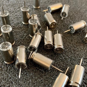 May include: A collection of silver bullet push pins. The bullet casings are hollow and have a clear plastic top with a sharp metal pin protruding from the top.