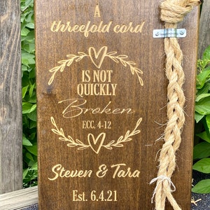 May include: A wooden sign with the text "A threefold cord is not quickly broken ECC. 4-12" and the names "Steven & Tara" with the date "Est. 6.4.21". A thick, braided rope is attached to the sign.