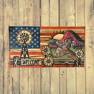 Könnte beinhalten: Holzschild mit amerikanischer Flagge, Windmühle und dem Wort "WELCOME". Die rechte Seite zeigt eine rote Scheune, einen Fasan, Sonnenblumen und eine untergehende Sonne. Das Schild ist in Rot, Blau, Braun und Gelb bemalt.