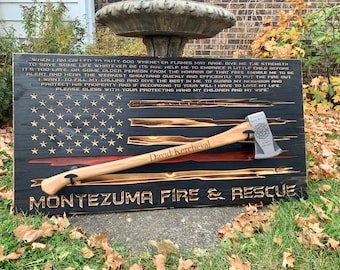 May include: A wooden sign with a black and red American flag design. The sign has the text "MONTEZUMA FIRE & RESCUE" and a firefighter's oath. A firefighter's axe is mounted on the sign with the name "David Kercheval" engraved on the handle.