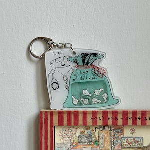 May include: A clear acrylic keychain featuring a cartoon doctor and a teal bag with the text "a bag of chill vibe." The bag contains several white thumbs-up icons. The keychain has a silver ring.