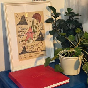 May include: A framed artwork with a whimsical illustration and the text "You thought you had it all figured out." A red book with the word "love" is on a blue surface. A potted plant is also visible.