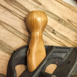May include: A wooden hand plane handle with a smooth, light brown finish. The handle is attached to a black metal hand plane.