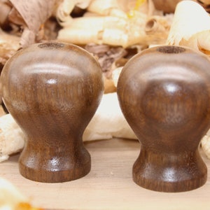May include: Two dark brown wooden knobs with a smooth, polished finish. The knobs are round with a slightly tapered base. They are sitting on a light brown wooden surface.