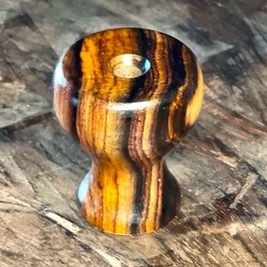 May include: A wooden bottle stopper with a rounded top and a flared base. The stopper is made of wood with a striped pattern of brown and yellow-orange tones. A hole is drilled in the center of the top.