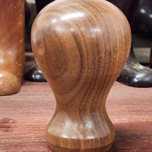 May include: A close-up of a wooden bottle stopper with a smooth, rounded top and a slightly flared base. The wood has a rich brown color and a beautiful grain pattern.