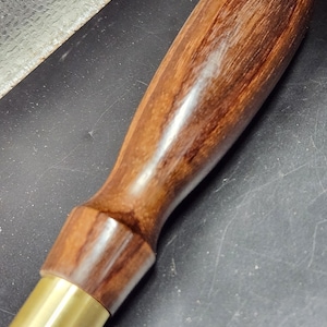May include: A handcrafted wooden handle with a brass ferrule. The handle is made of dark brown wood with visible grain, and the brass ferrule is a bright gold color. The handle is likely for a tool or instrument.
