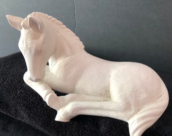 May include: White ceramic figurine of a horse lying down with its head resting on its front leg. The horse is in a relaxed pose and has a smooth, matte finish.
