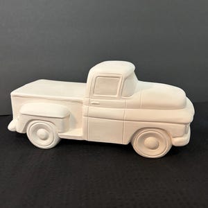 May include: A white ceramic pickup truck figurine. The truck has a classic design with a cab, bed, and wheels. The figurine is unpainted and ready for customization. The truck is a decorative item.