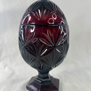 Puede incluir: Un platito de caramelo de vidrio rojo con una tapa en forma de huevo. El plato tiene un diseño de vidrio cortado y se asienta sobre una base hexagonal.