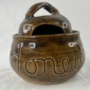 Könnte beinhalten: Ein brauner Keramik-Honigtopf mit Deckel und Henkel. Das Wort "Honey" ist auf der Vorderseite des Topfes eingeprägt.