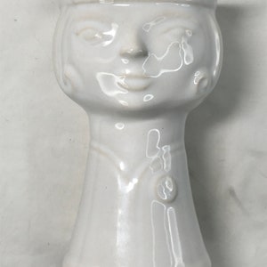 Puede incluir: Maceta de cerámica blanca con forma de cabeza humana con una cara sonriente simple. La maceta tiene una abertura ancha en la parte superior y una base estrecha.