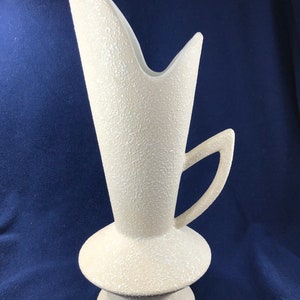 May include: A white ceramic vase with a textured surface and a unique, angular design. The vase has a tall, slender body with a wide, flared opening and a handle on one side. The base of the vase is round and has a textured surface.