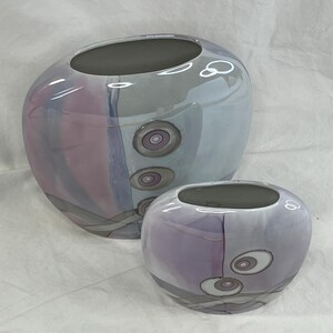 May include: Two ceramic vases with a light purple, pink, and gray abstract design. The larger vase has three concentric circles with a pink center and gray outer ring. The smaller vase has two concentric circles with a white center and gray outer ring.