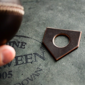 May include: A dark brown leather patch with a circular hole in the center. The patch has a rounded, pointed shape and is on a green textured surface. The text "Genuine" and "1905" are visible in the background.