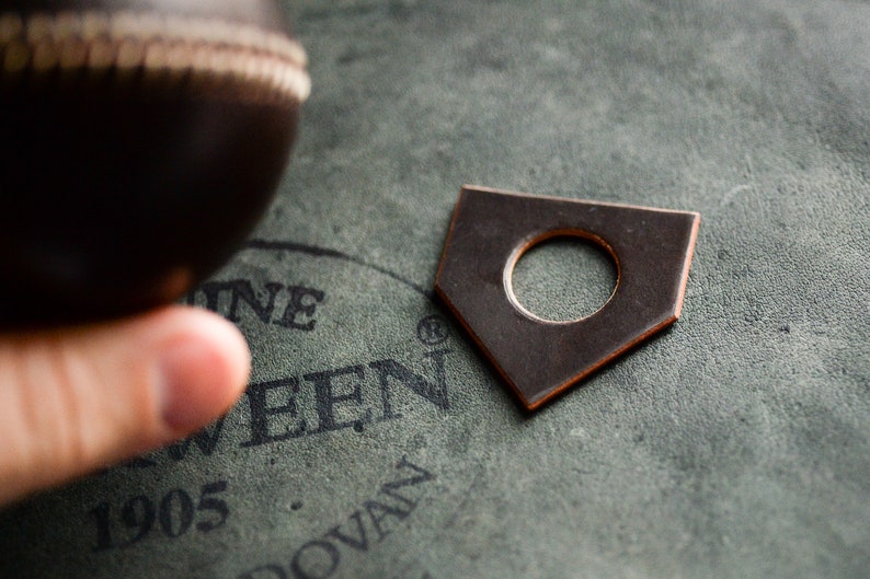 May include: A dark brown leather patch with a circular hole in the center. The patch has a rounded, pointed shape and is on a green textured surface. The text "Genuine" and "1905" are visible in the background.