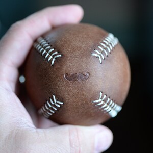 May include: A brown leather baseball with white stitching and a mustache design.