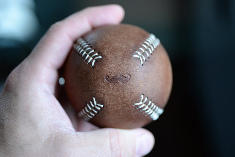 May include: A brown leather baseball with white stitching and a mustache design.