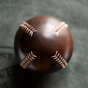 May include: A brown leather baseball with white stitching. The baseball is on a gray surface.