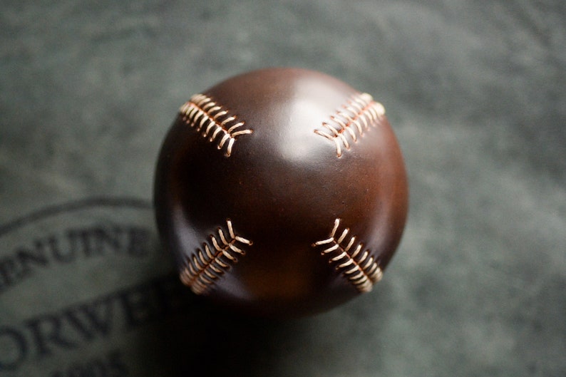 May include: A brown leather baseball with white stitching. The baseball is on a gray surface.