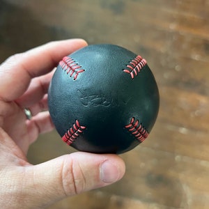 May include: A black leather baseball with red stitching. The baseball has the word "Truly" embossed on it.