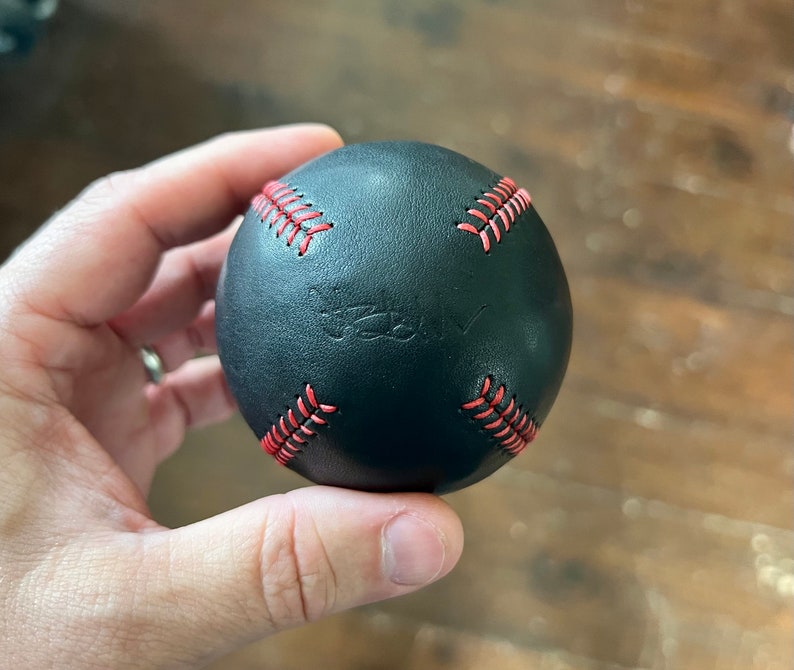 May include: A black leather baseball with red stitching. The baseball has the word "Truly" embossed on it.