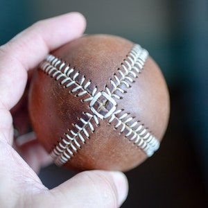 May include: A brown leather baseball with white stitching. The ball is being held in a hand.