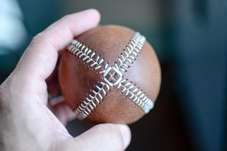 May include: A brown leather baseball with white stitching. The ball is being held in a hand.