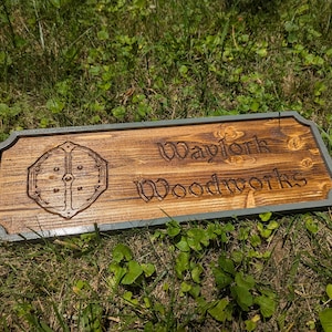 May include: A rectangular wooden sign with a shield design and the text "Waylork Woodworks" engraved on it. The sign has a dark brown finish with a grey border, set against a backdrop of green grass.