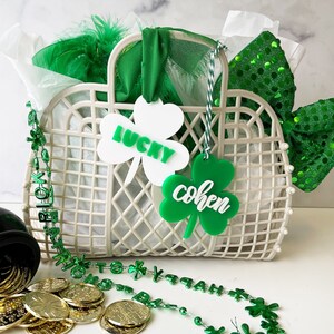 Pu&ograve; includere: Un cesto di vimini bianco con etichette a forma di trifoglio verde che recitano "Lucky" e "Cohen". Il cesto &egrave; pieno di carta velina verde e bianca, una boa di piume verde e bianca e un fiocco verde con paillettes. Davanti al cesto c'&egrave; una collana di perline a forma di trifoglio verde.