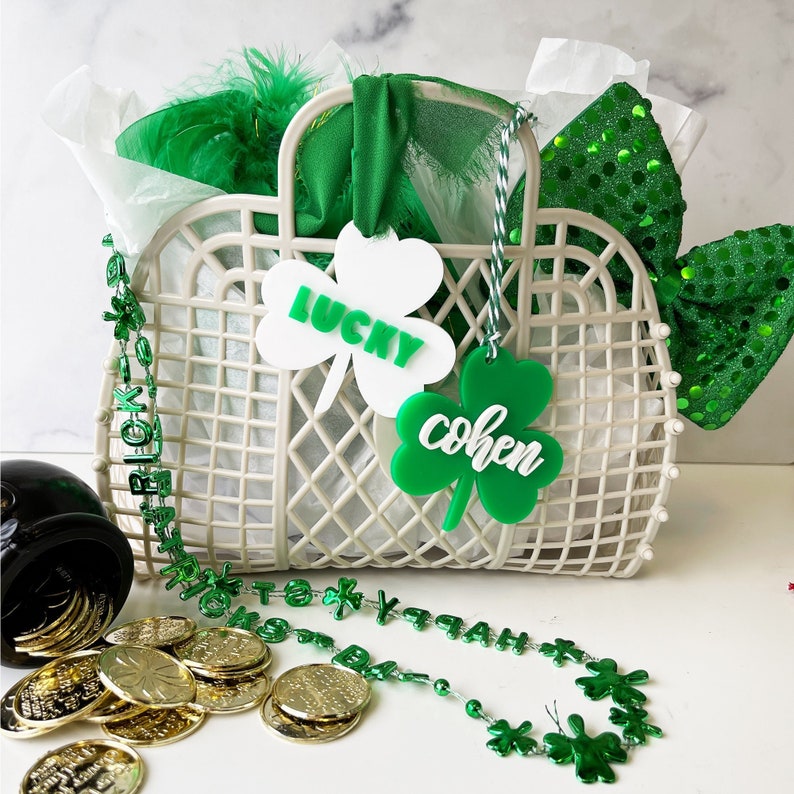 Pu&ograve; includere: Un cesto di vimini bianco con etichette a forma di trifoglio verde che recitano "Lucky" e "Cohen". Il cesto &egrave; pieno di carta velina verde e bianca, una boa di piume verde e bianca e un fiocco verde con paillettes. Davanti al cesto c'&egrave; una collana di perline a forma di trifoglio verde.